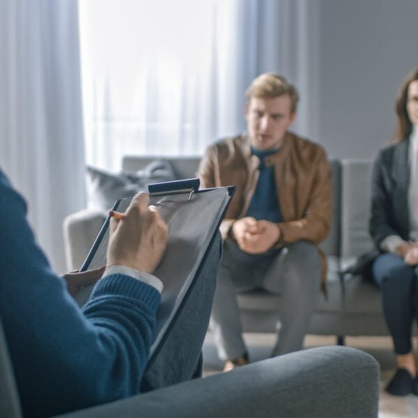 Couple on a Counseling Session with Psychotherapist. Foreground Focus on Back of the Therapist Taking Notes and Talking: People Sitting on the Analyst Couch, Discussing Psychological Problems